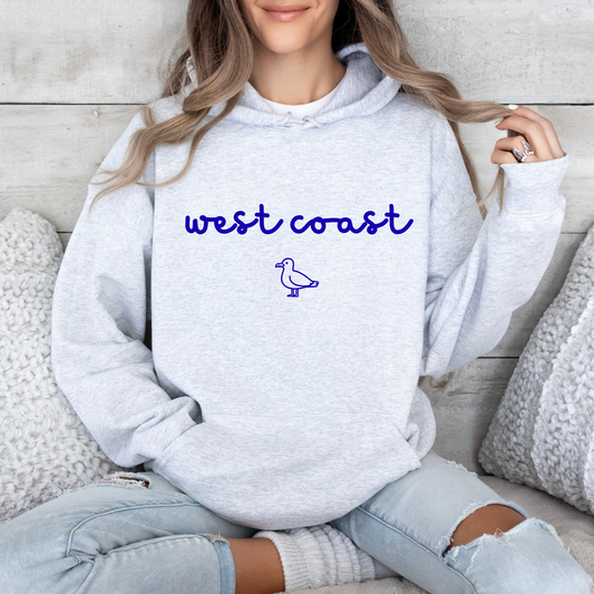 A woman wearing a light heather gray hoodie with 'west coast' and a small bird graphic printed in blue on the chest, sitting on a couch with pillows.