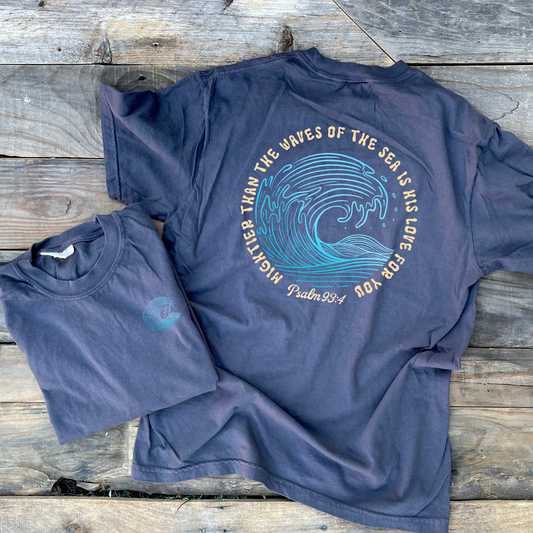A gray t-shirt is laid flat on a rustic wooden plank surface, showing the back of the shirt with a large graphic of a blue ocean wave and the text 'MIGHTIER THAN THE WAVES OF THE SEA IS LOVE FOR YOU Psalm 104:4'. A smaller folded view of the front or inner side is placed at the lower left.