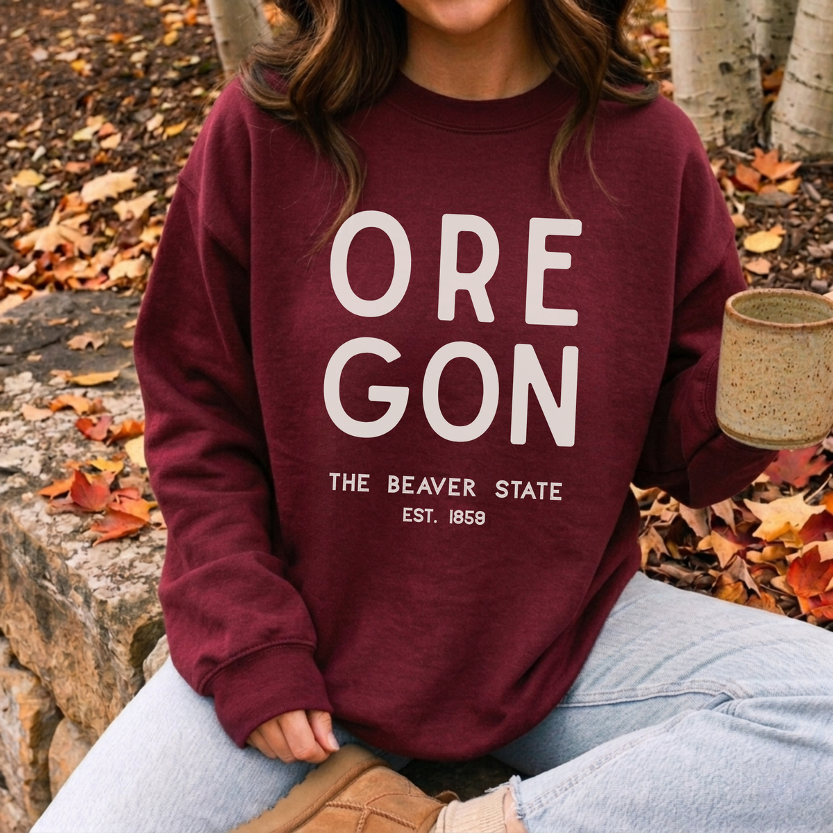 A person wearing a burgundy crewneck sweatshirt with 'OREGON THE BEAVER STATE EST. 1859' printed in white on the front, sitting outdoors on a stone bench surrounded by fallen autumn leaves and holding a ceramic mug.