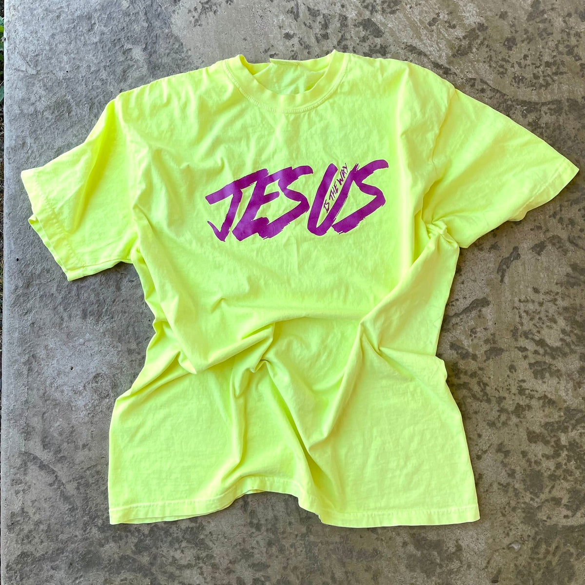 A neon yellow-green t-shirt with the word 'JESUS' printed across the chest in purple script, laid flat on a textured concrete floor.