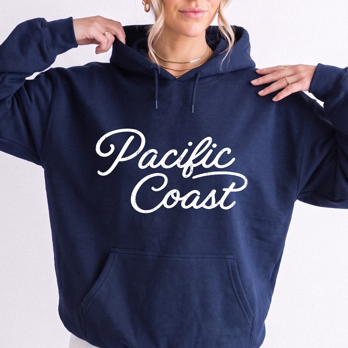 A woman wearing a navy blue hoodie with 'Pacific Coast' printed in white script across the chest, posed against a plain white background.