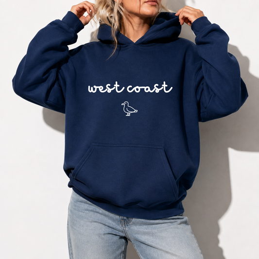 A woman wearing a navy blue hoodie with 'west coast' and a small bird outline printed in white on the chest, paired with light-wash jeans against a white wall.