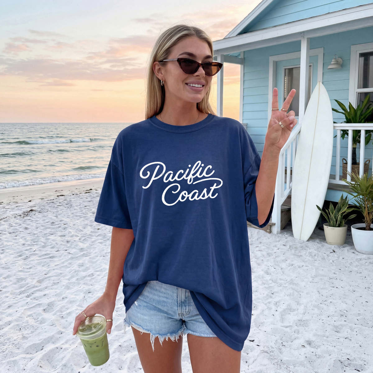 A woman stands on a sandy beach wearing an oversized navy blue t-shirt with 'Pacific Coast' printed in white script across the chest. She is also wearing denim shorts, sunglasses, and holding an iced green drink, with a light blue beach house and surfboards in the background.