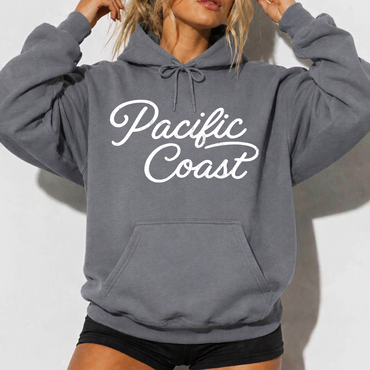 A woman wearing a dark gray pullover hoodie with 'Pacific Coast' printed in white script across the chest, paired with black shorts against a light gray wall.