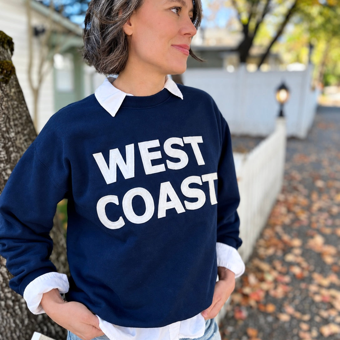 A woman wearing a navy blue crewneck sweatshirt with 'WEST COAST' printed in bold white letters across the chest, layered over a white collared shirt and light blue jeans, standing outdoors.