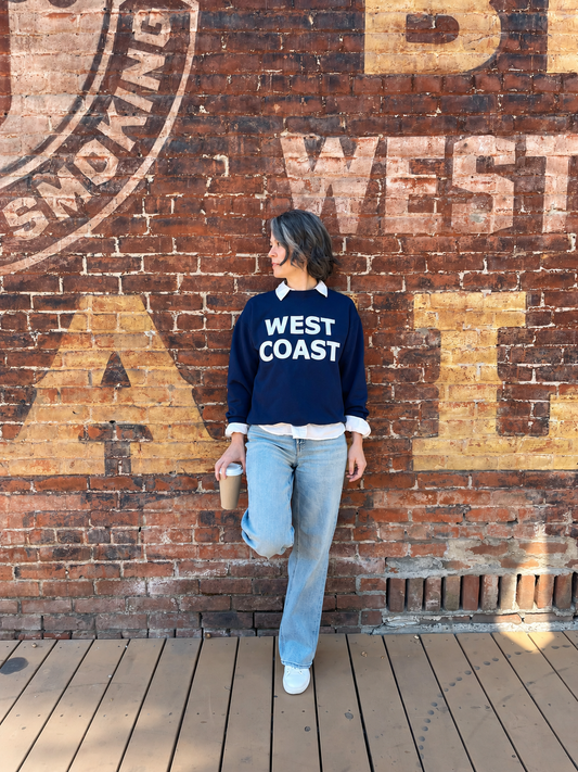 A woman stands on a wooden deck in front of a brick wall with large painted letters, wearing a navy blue sweatshirt with 'WEST COAST' printed in white across the chest, light blue jeans, and white sneakers, while holding a coffee cup.